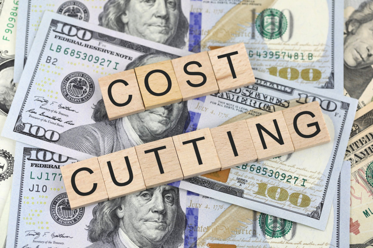 A picture of wooden tiles with letters stamped on spelling cost cutting lying on top of a pile of dollar bills.