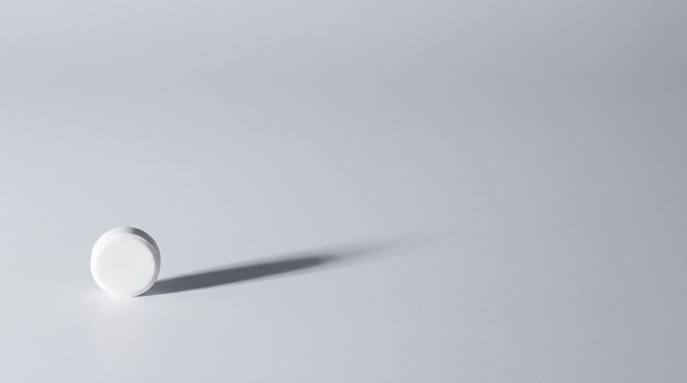 One small white pill casting a long shadow across an empty grey surface, illustrating how a single case becomes a statistic