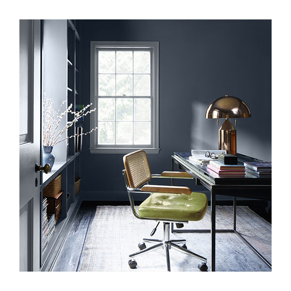 A navy blue painted home office with a rectangular desk, a gold domed desk lamp, and a rolling office chair with a lime green seat.