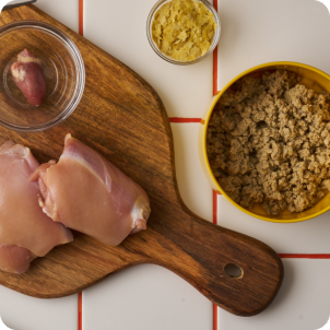 Overhead photo of raw chicken and other whole-food ingredients arranged on a surface with wooden scoops